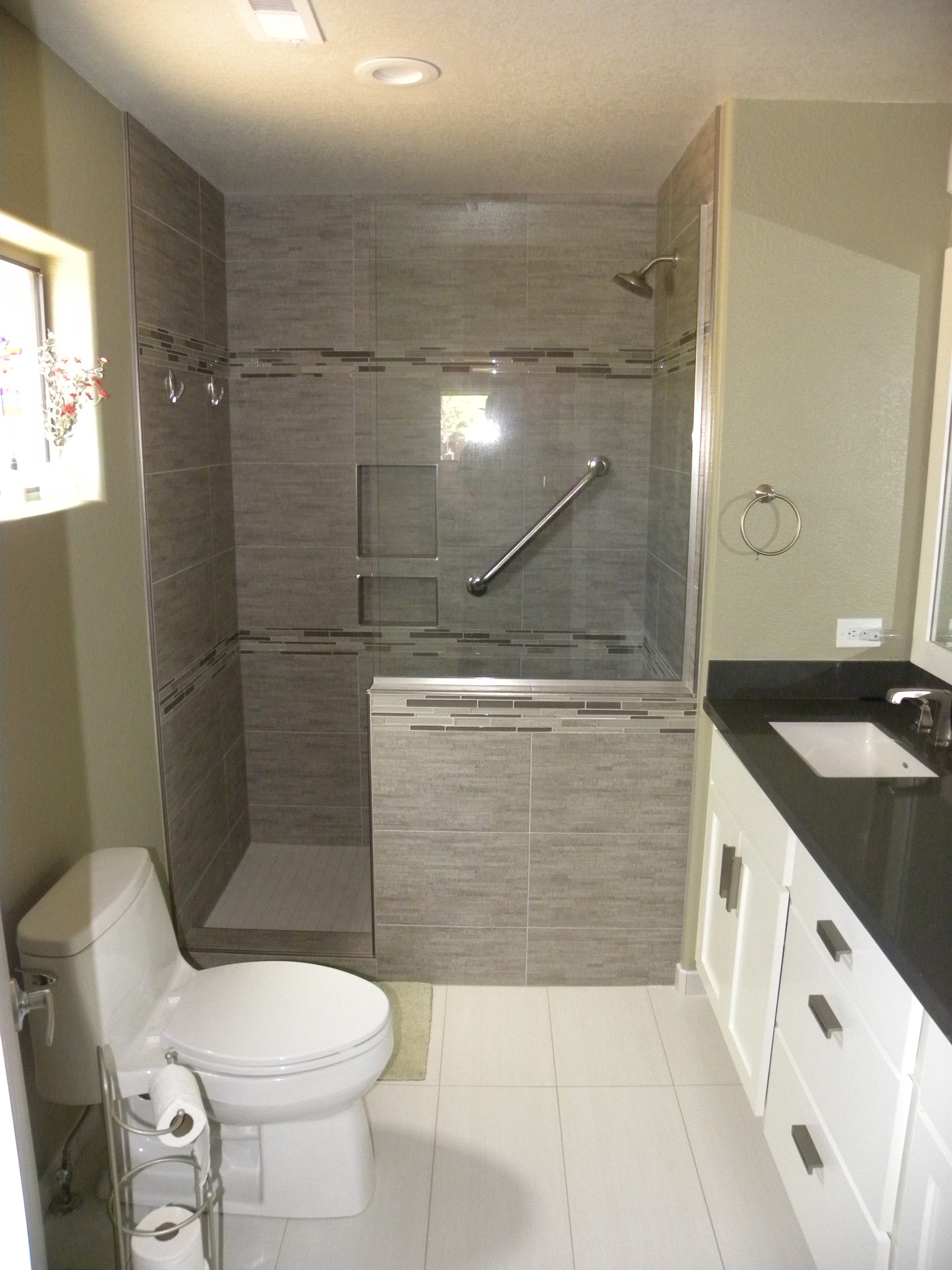After photo of a custom residential bathroom with black countertops and white wooden cabinets, new white tiles, and a custom designed shower with gray tile, glass windows and doors, and specially design handles.